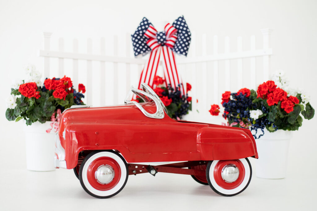 Patriotic summer photo studio set featuring red, white, and blue flowers, white picket fence, and vintage red car in Jacksonville, FL.