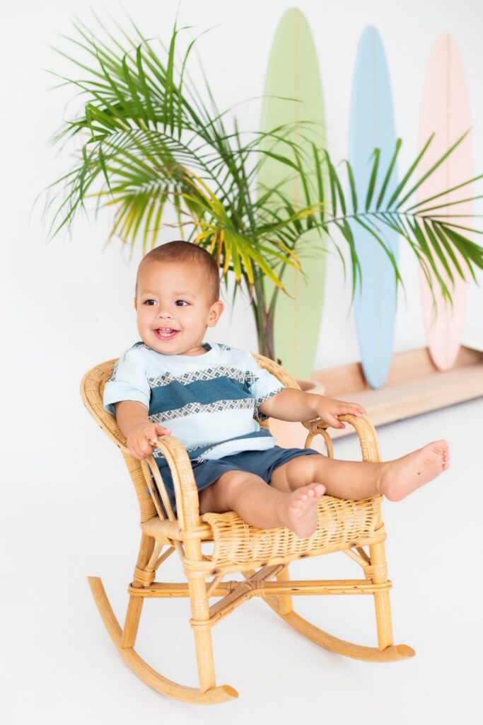 Young boy sitting in a wicker rocking chair in front of palm tree and surfboard set at Jacksonville photo studio.
