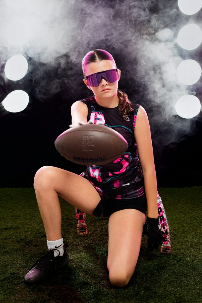 Indoor Jacksonville sports photography session featuring athlete kneeling on turf holding football with dramatic fog and stadium lighting around her.