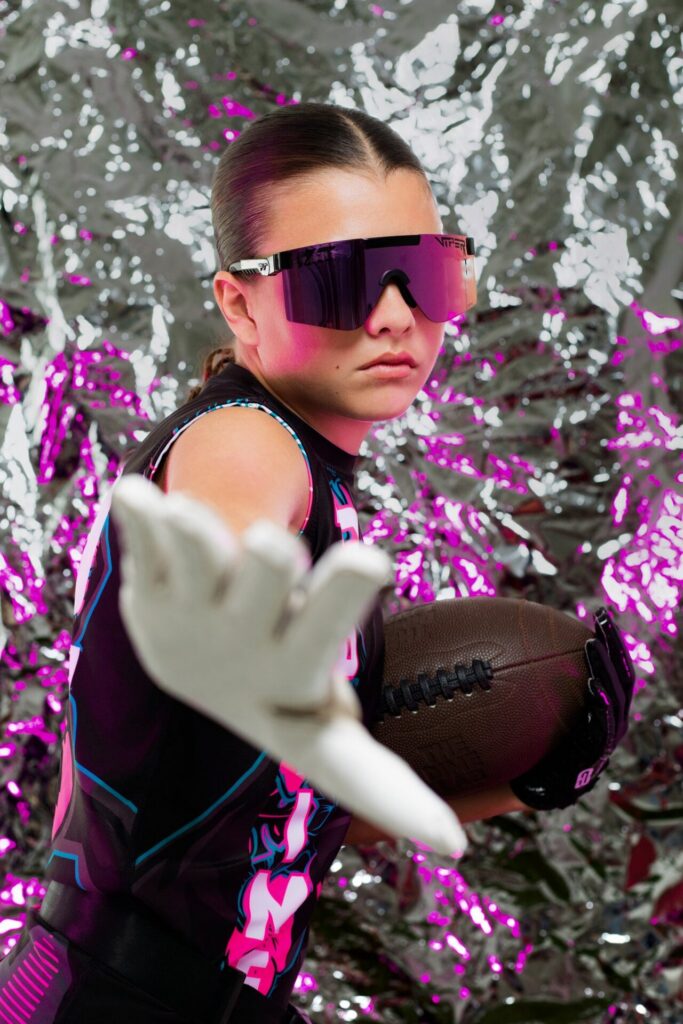Flag football athlete holding football with hand blocking camera during dramatic sports photography session at Jacksonville photo studio with foil backdrop.