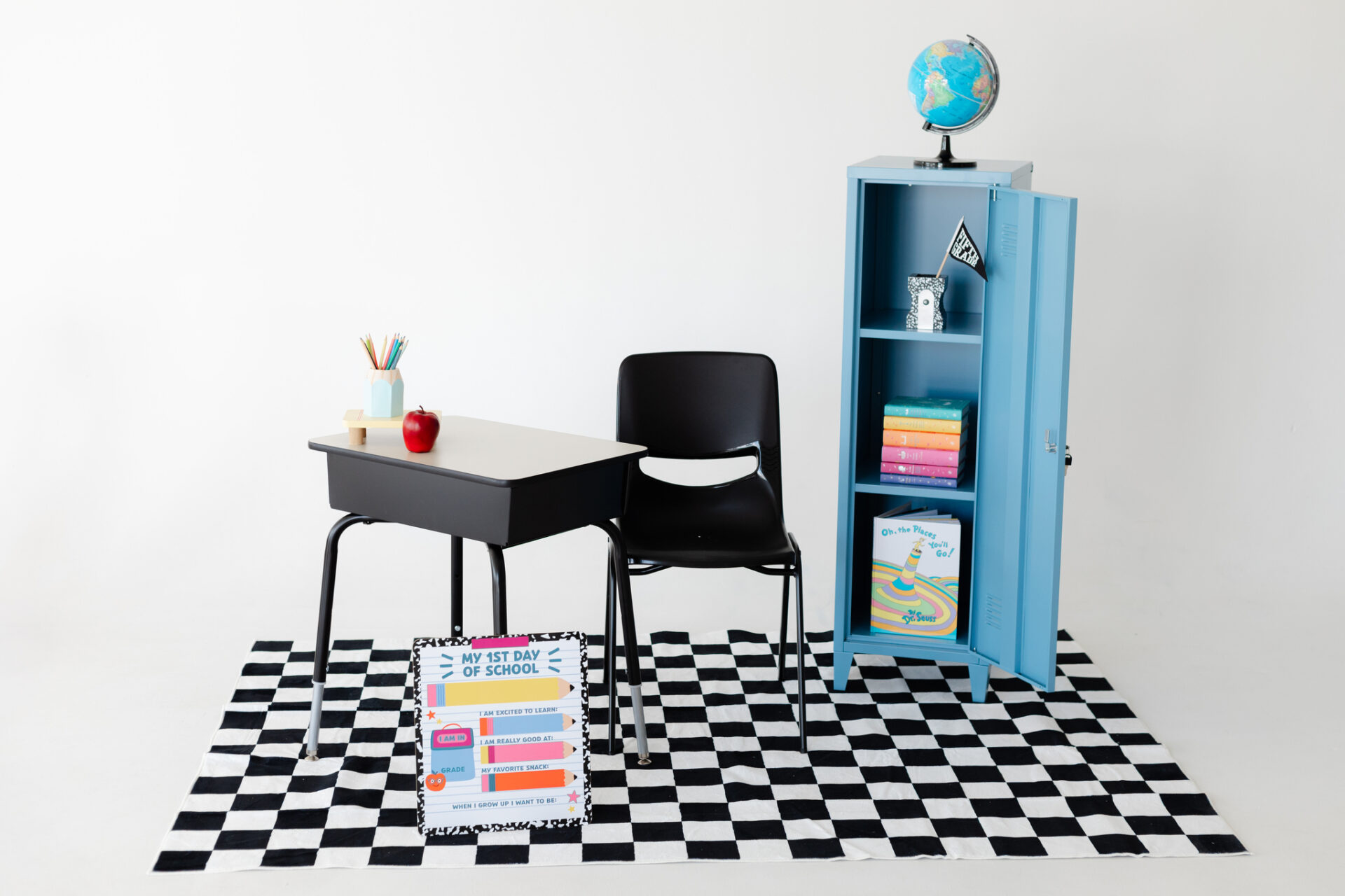 A classic black and metal school desk over a checkerboard rug with a blue locker in a white photo studio