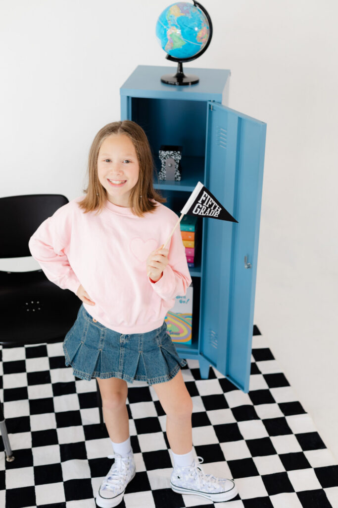5th grade girl in a pink sweatshirt holding a fifth grade pennant and posing for her back to school mini session
