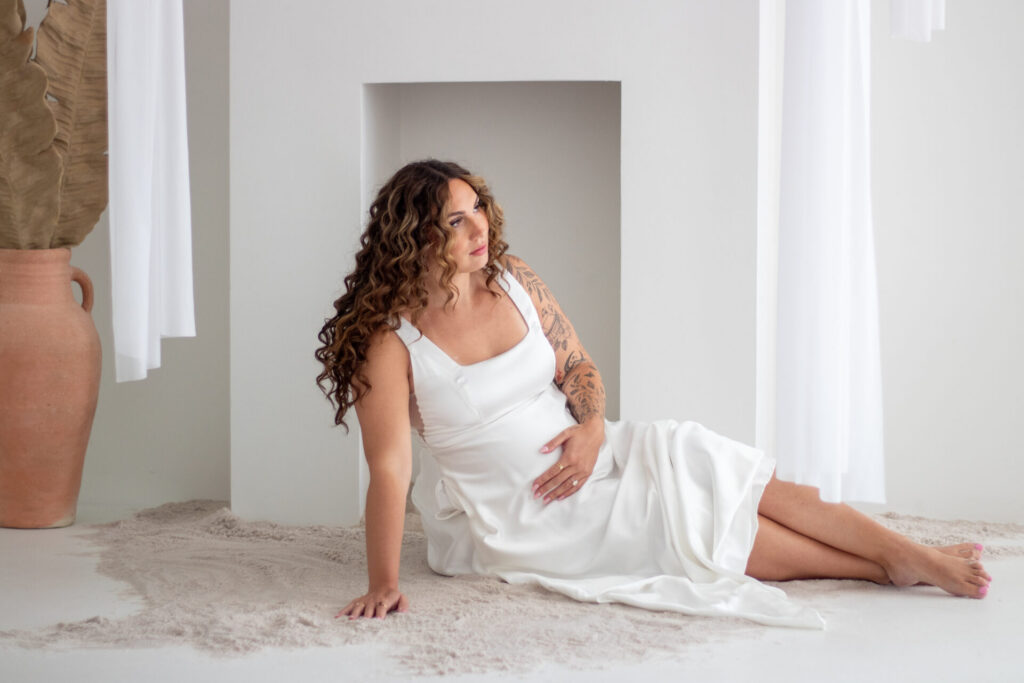 Maternity session photographed by laylagphotos, mother sitting in front of a beach-inspired fireplace with sand, in a satin white dress, holding her belly and looking away from the camera.