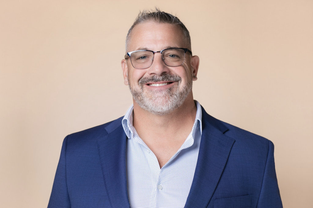 Professional headshot of a man wearing glasses and a navy blue suit against a tan neutral backdrop