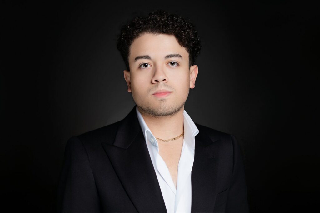 Professional headshot of young man in a tailored shirt and jacket on a black background with a serious facial expression.