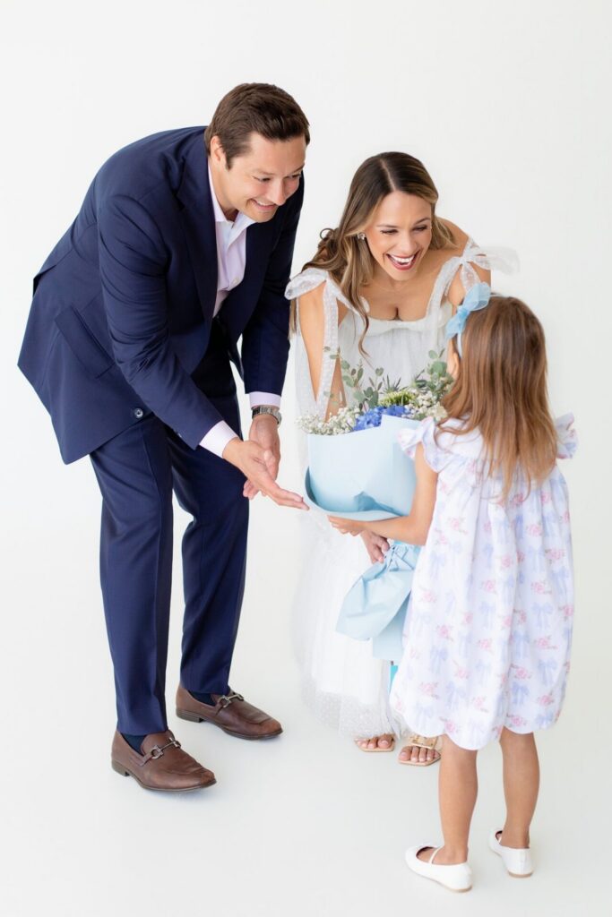 Daughter handing blue gender reveal flower bouquet to her smiling parents in all white natural light photo studio in Jacksonville.