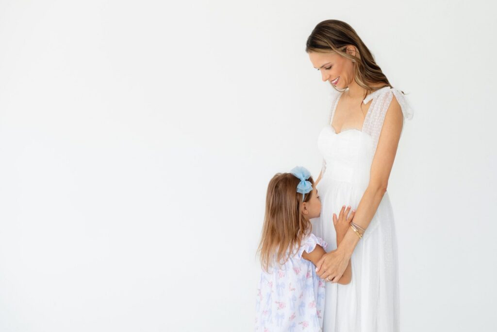 Young daughter kisses her mom's belly during pregnancy announcement and gender reveal photoshoot in Jacksonville, FL photo studio.