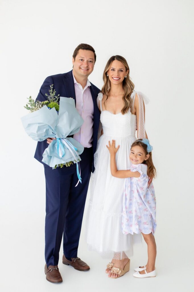 Family portrait at a Jacksonville photo studio after a gender reveal, with dad holding a blue flower bouquet and big sister touching mom's pregnant belly.