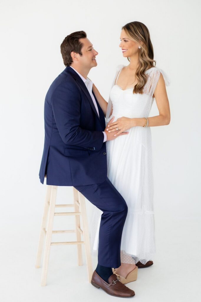 Husband sits on a stool and holds wife's pregnant belly in a couple's portrait session for pregnancy announcement in a photo studio.